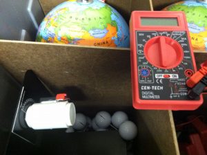 A red digital multimeter is placed on top of a wooden box. It is connected to a white cylindrical object with red and black wires. A globe labeled with Russia, Kazakhstan, Mongolia, and China is visible in the background.