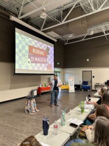 A male presenter in a light blue sport coat stands and holds a book in front of a large screen displaying the graphic "Reading is Magical" in a large conference room.