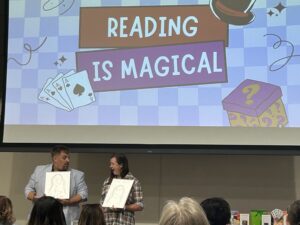 Two presenters, a man and a woman, stand in front of a screen displaying the graphic "Reading is Magical." They are holding up simple portrait drawings and looking at each other.