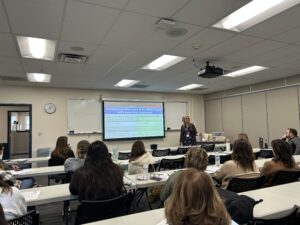 A female presenter stands next to a projected screen that reads "Promoting Relevance & Building Depth with Essential Questions" to a room of seated educators during a training session.