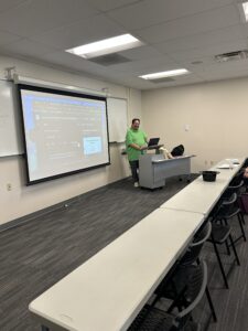 A male presenter in a green shirt stands at a podium next to a large screen displaying an online collaboration tool to an audience seated in a classroom setting.