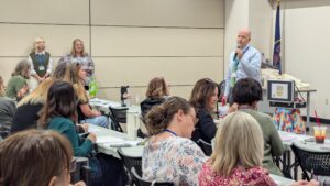 A male presenter in a blue shirt speaks into a handheld microphone to a group of seated educators during a professional development session. Two women stand behind the audience near the Utah state flag.