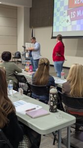 A male presenter in a light blue shirt holds a book and speaks to an audience of seated educators during a session. A woman in a red sweater stands next to him.