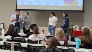 A a man in a blue shirt and light blue jacket, and three women in casual dress stand facing an audience seated at the SUMS conference. Above them, a screen displays a graphic with the word "MAGICAL," playing cards, and a question mark box, which is part of the "Reading is Magical" theme.