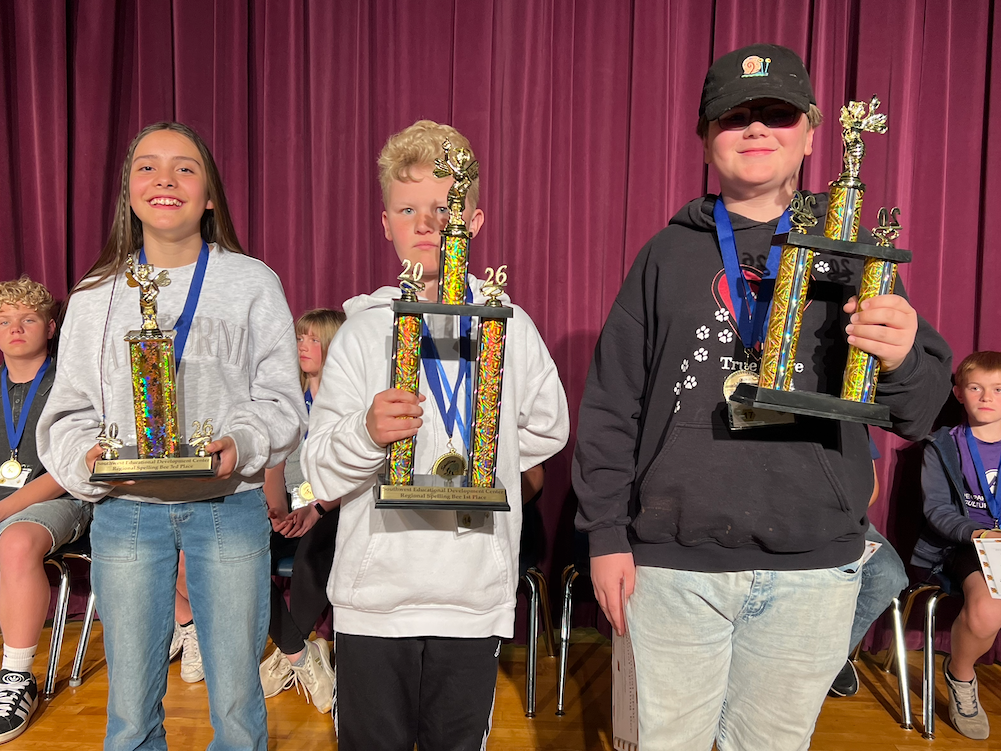 Three students stand proudly on a stage with large trophies at the 8th Annual SEDC Regional Spelling Bee in front of a purple curtain.