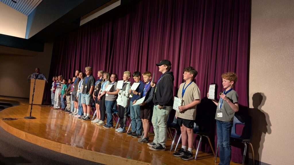 A group of 18 students stand in a line on a stage, holding certificates and medals, at the 8th Annual SEDC Regional Spelling Bee.